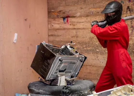 Person in red protective suit and helmet swinging bat at electronic equipment in rage room.
