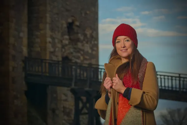 Woman in red hat and brown coat standing in front of historic stone tower and bridge.