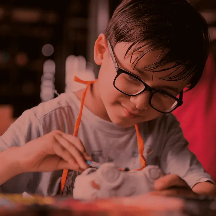 Child wearing glasses concentrates on painting pottery at a table under warm lighting.