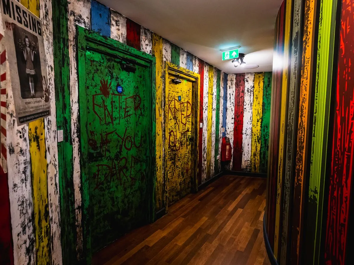 A corridor with brightly coloured distressed doors in green, yellow and red, covered with graffiti-style markings, leading to an escape room experience.