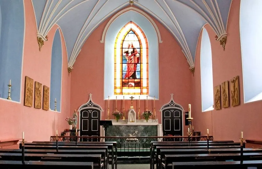 Interior of a pink-walled chapel with a stained glass window above the altar and wooden pews in the foreground.