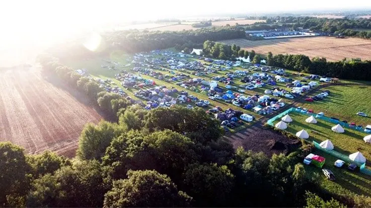 Aerial view of a large campsite with hundreds of tents and vehicles arranged in rows across green fields at sunrise.