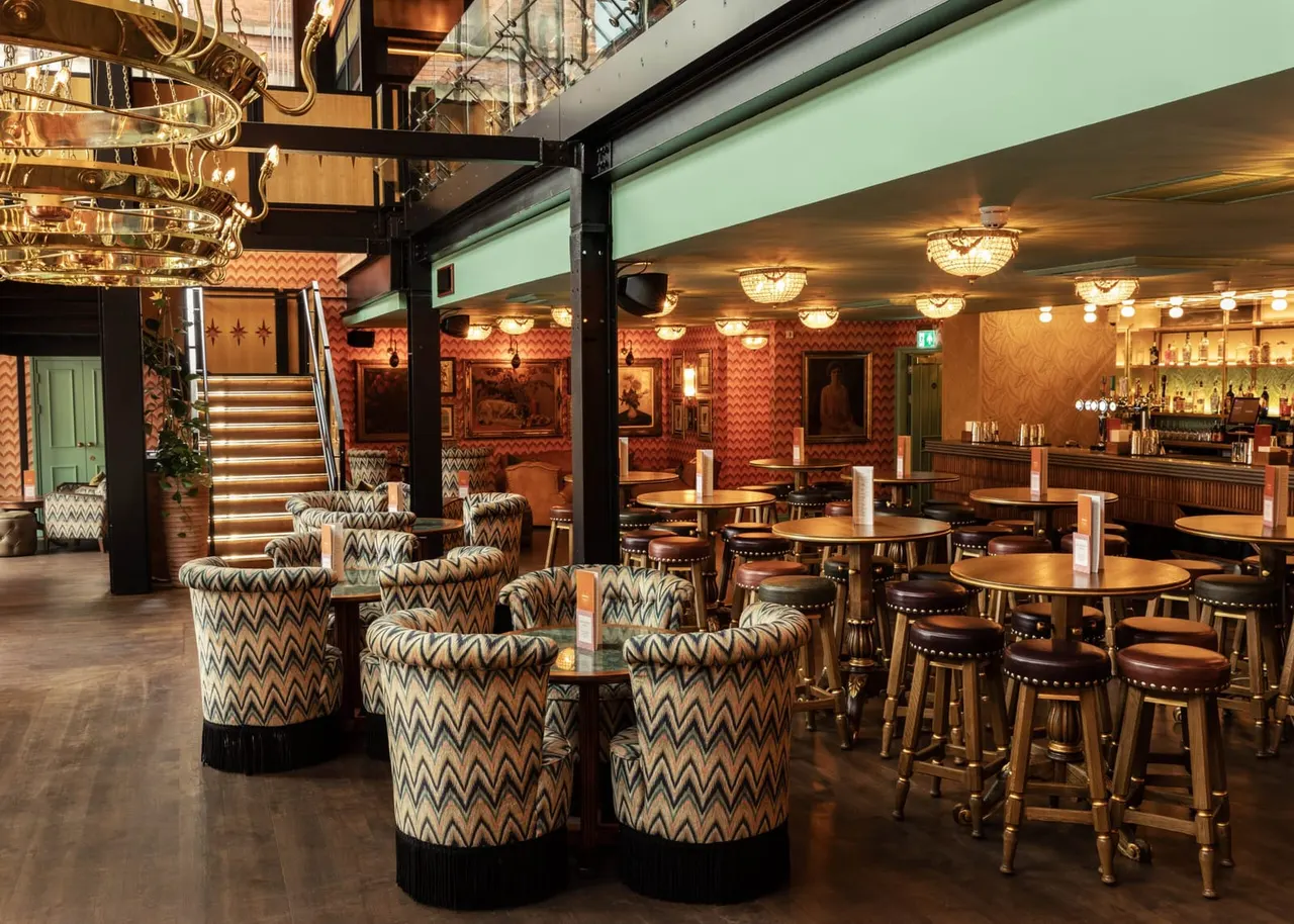 Art deco-style bar and lounge with patterned armchairs, dining tables, brass chandeliers and red damask wallpaper.