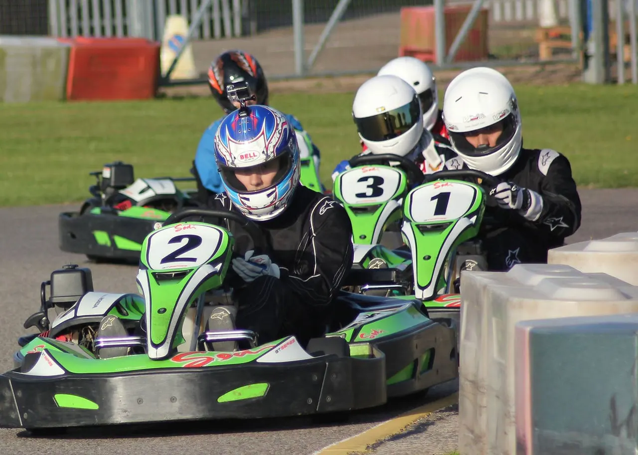 Go-kart racers in helmets navigating a track at a leisure venue, with numbered karts and safety barriers visible.