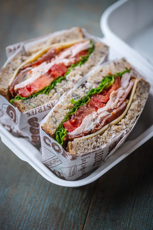 Two wholemeal sandwiches filled with bacon, lettuce, tomato and cheese in a white container.