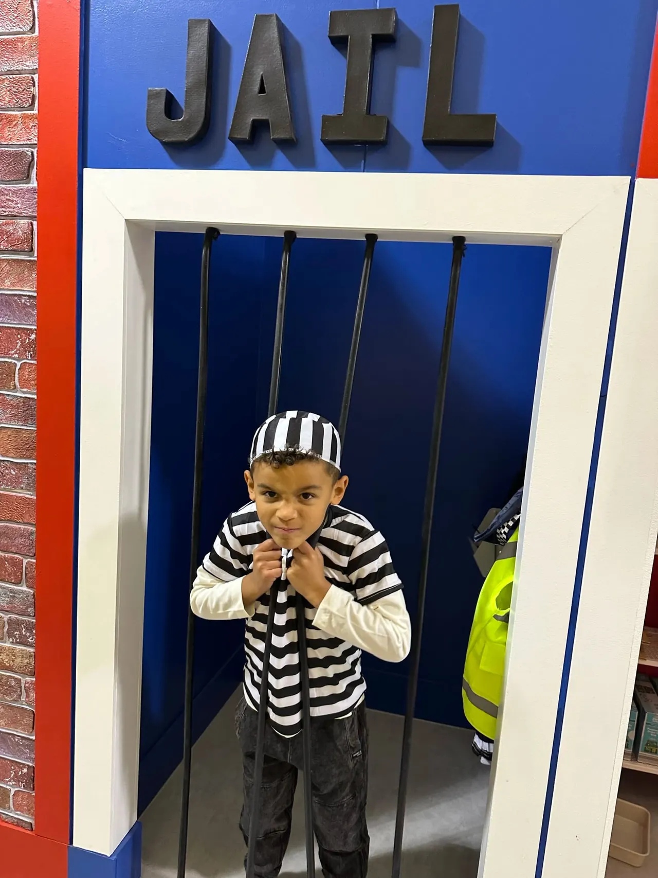 Young child in striped shirt and hat posing in jail cell display at leisure venue.