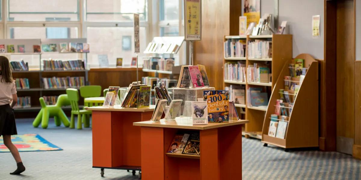 Children's library section with book displays, shelving, and bright green chair seating near large windows.
