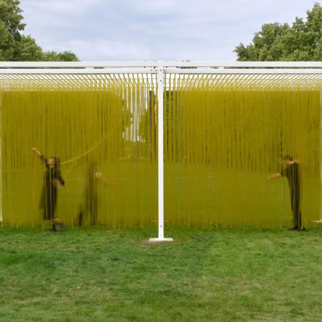 Two people in motion within a contemporary art installation with yellow vertical strings suspended between white frames in a grassy outdoor setting.
