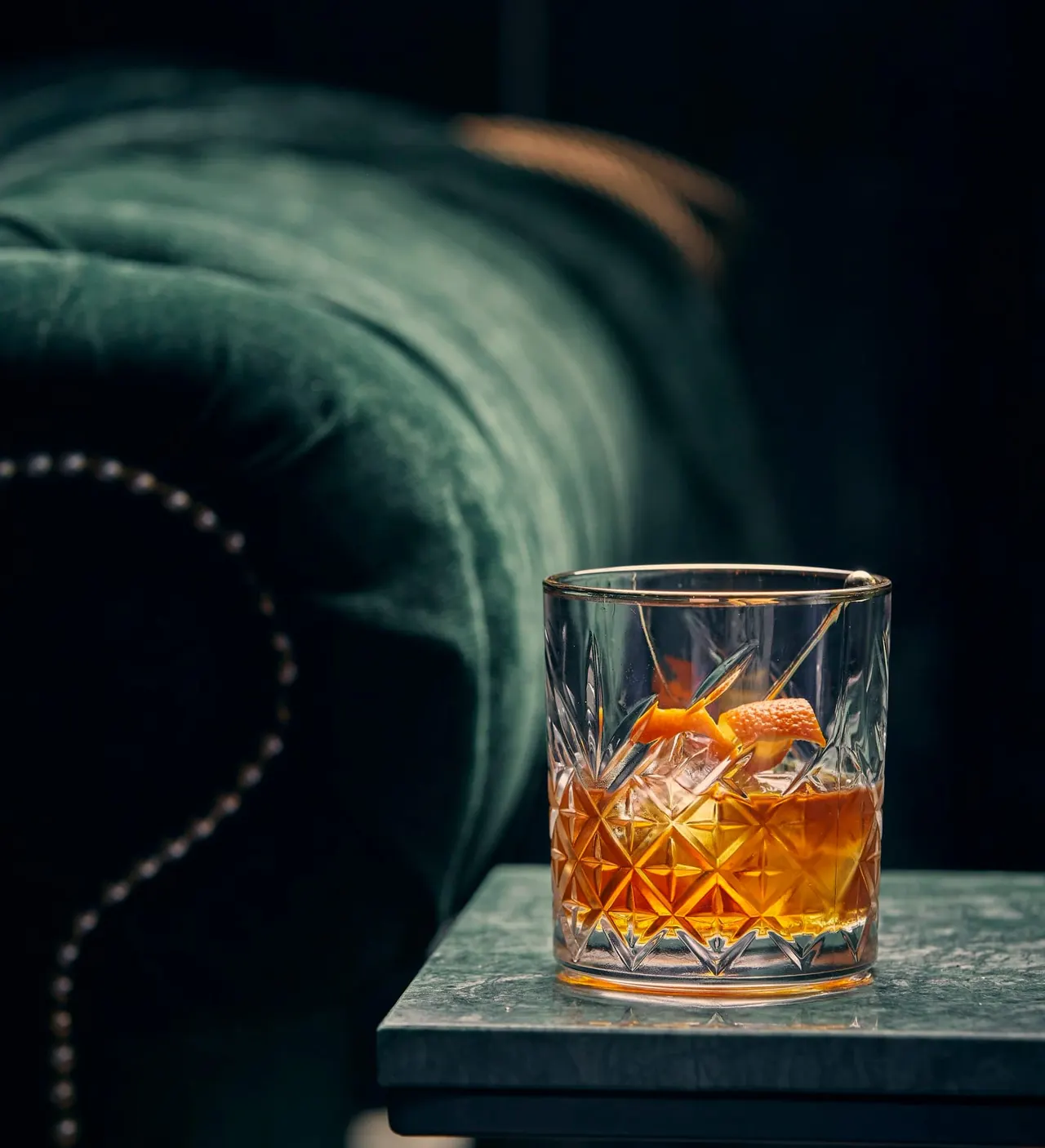 Crystal tumbler of whisky with ice on a dark surface, with a leather armchair blurred in the background.