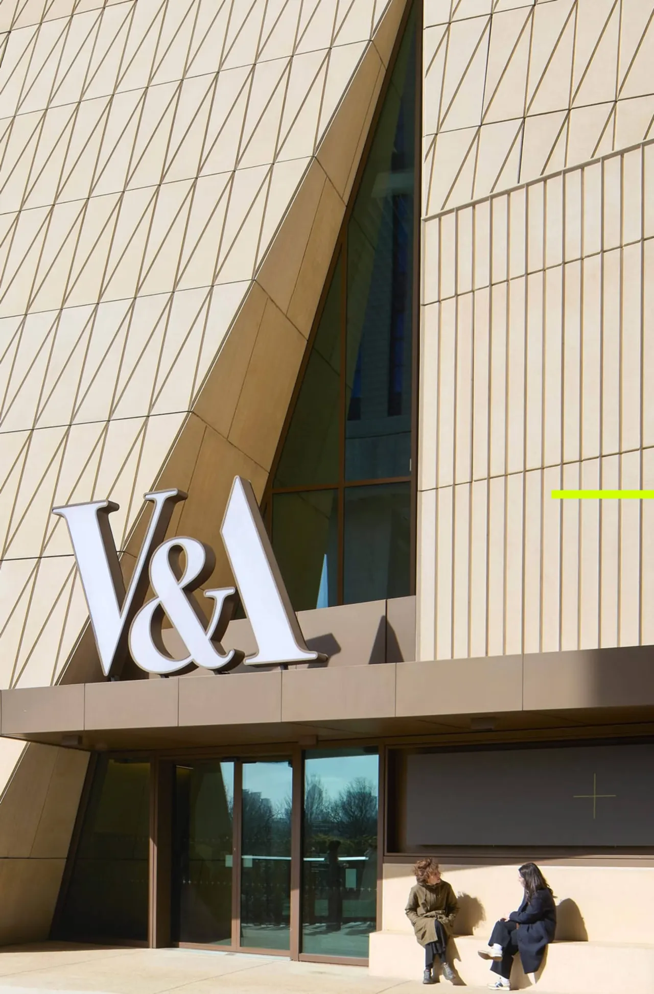 Two visitors stand outside a modern building with geometric tiled cladding and a large V&A sign.