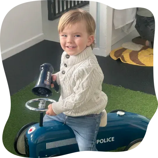 Toddler in cream knit jumper sits on blue toy police car indoors on artificial grass.