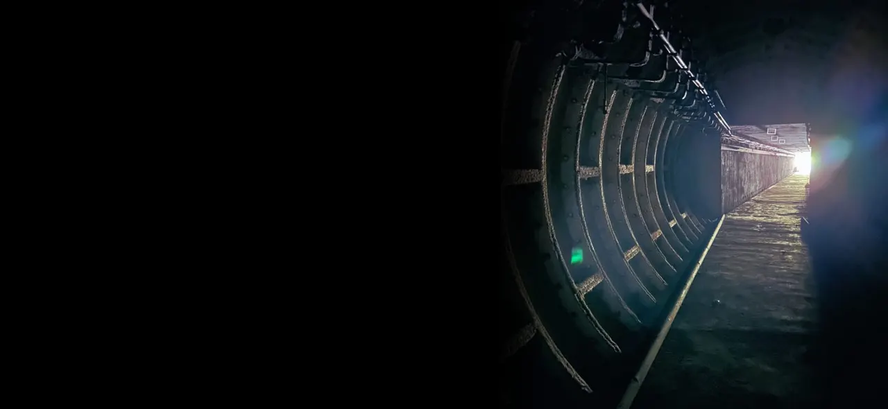 Interior of a dark underground tunnel with curved metal ribbed walls lit by a distant light source, showing the length of a fortified passage.