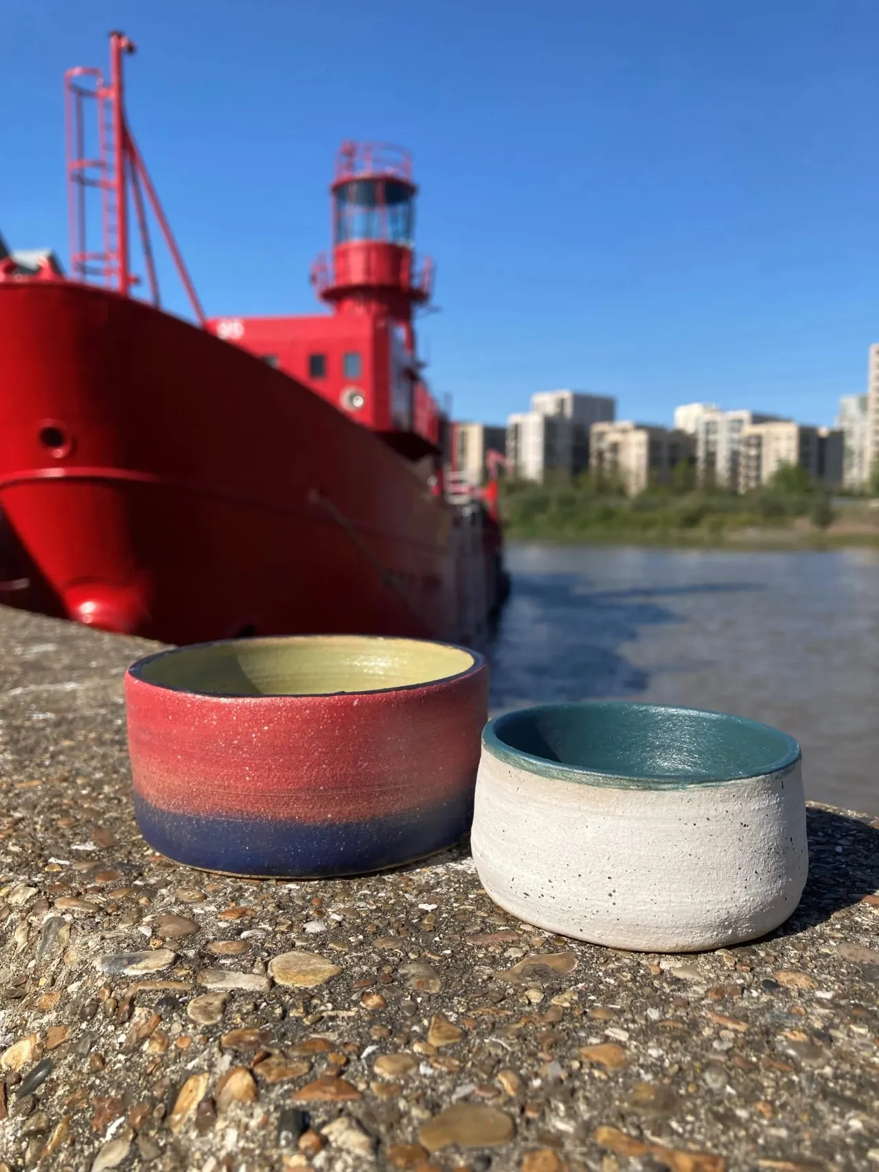 Two ceramic bowls on a waterside path with a red lighthouse and river behind.