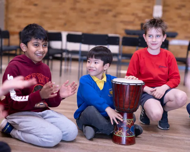 Three children sitting on a wooden floor playing a drum together in a school hall.