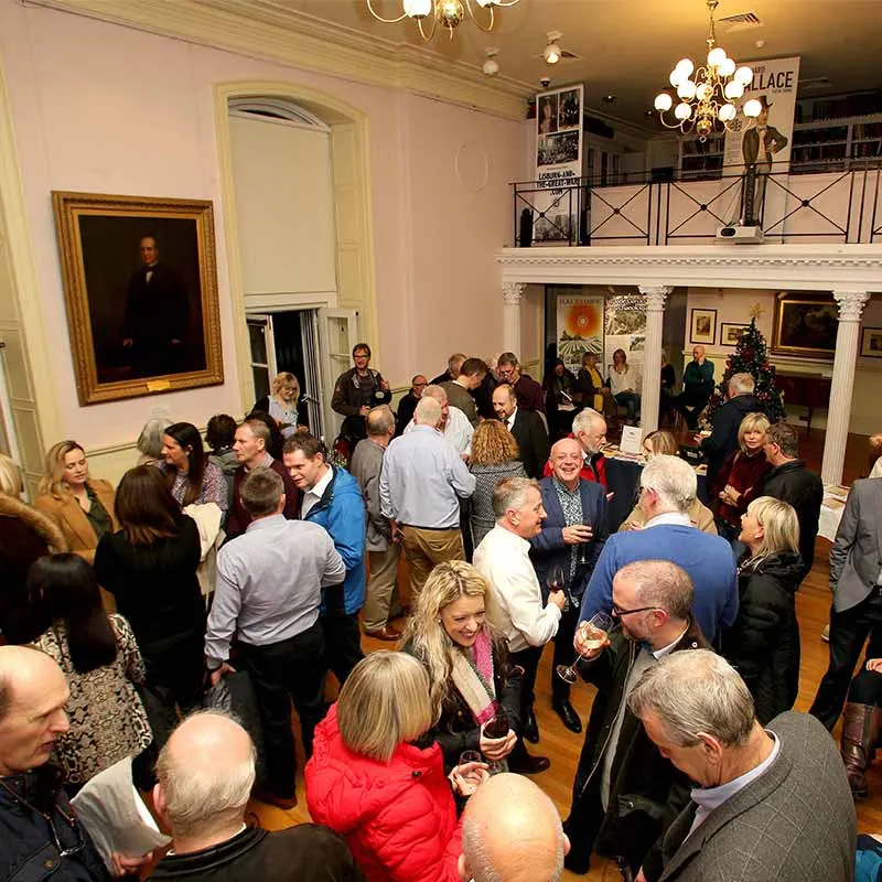 A crowd of visitors mingles and socialises in an elegant period room with a fireplace, chandelier and portrait painting.