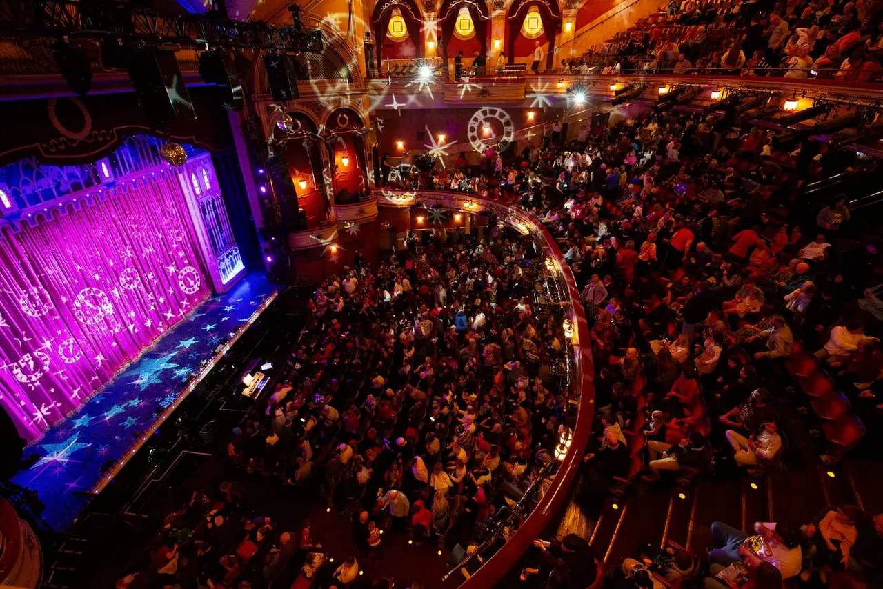 Overhead view of packed circus theatre with audiences seated around a ring lit by dramatic purple and gold lighting.
