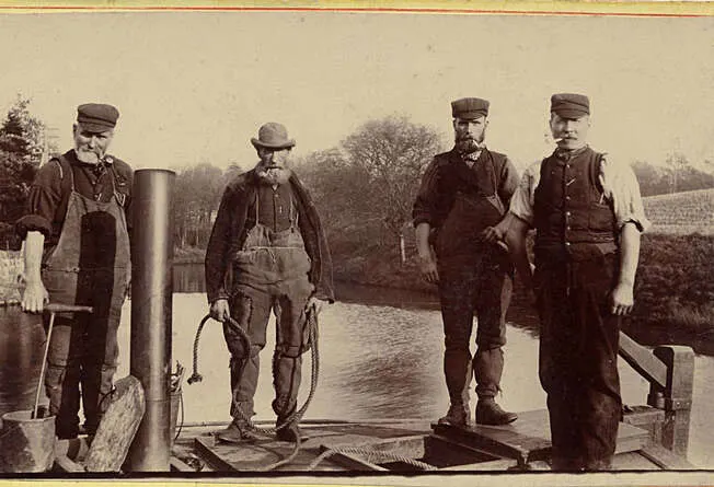 Four men in period work clothes and caps stand on a canal lock with machinery and tools beside a waterway.