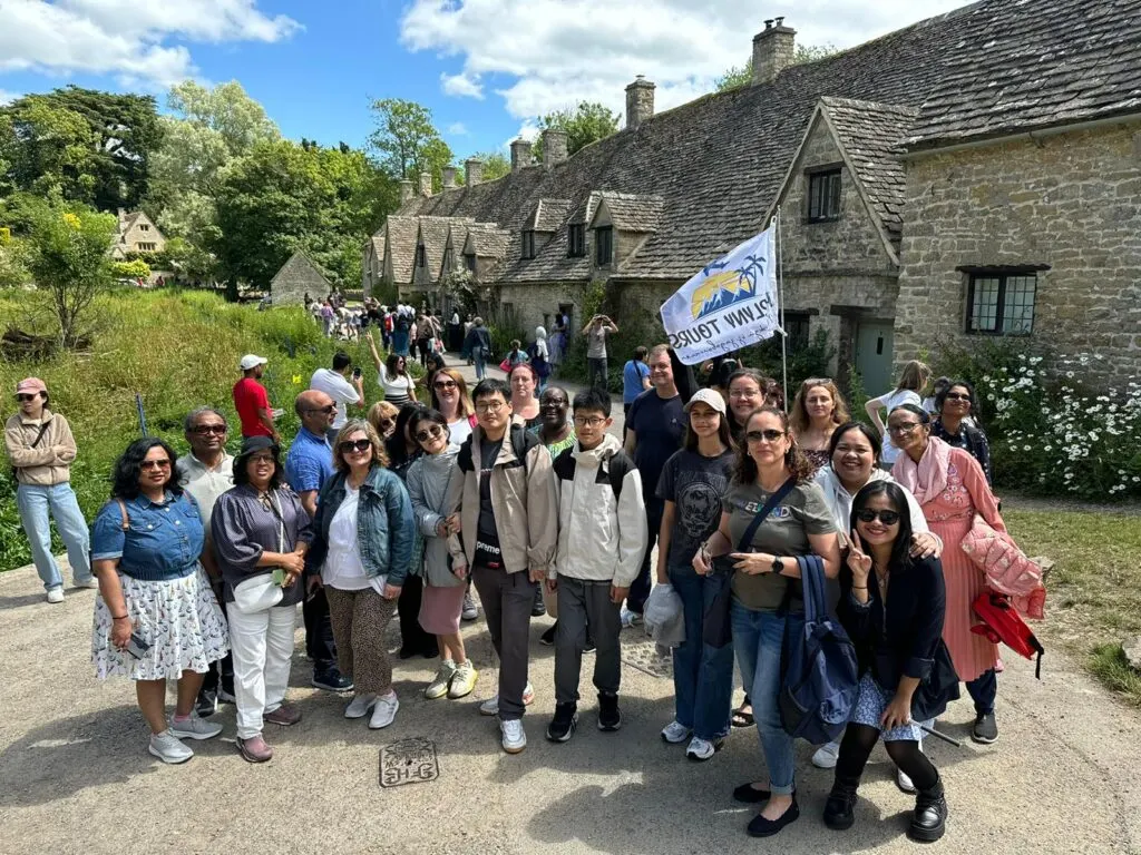 A large group of visitors pose together on a path in front of stone cottages, some holding a tour company flag.
