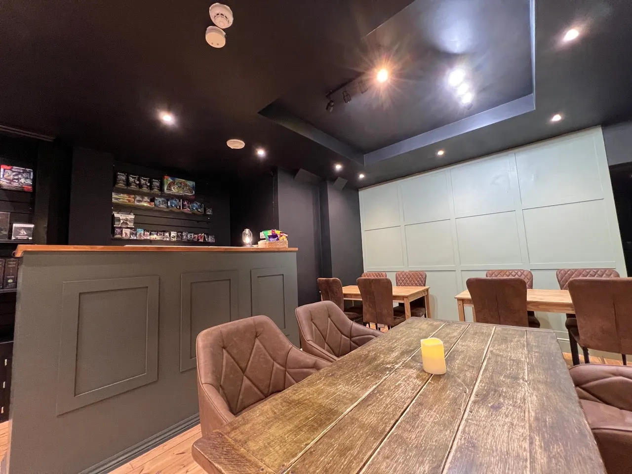Interior of a gaming venue with tables, brown upholstered chairs, a service counter, and shelving displaying board games and collectibles.