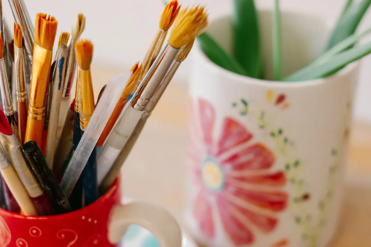 Paintbrushes in a red holder next to a painted ceramic pot with a flower design and potted plant.