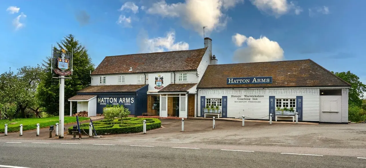 Historic white-painted coaching inn with dark roof beside a canal, with signage and pub sign under blue sky.