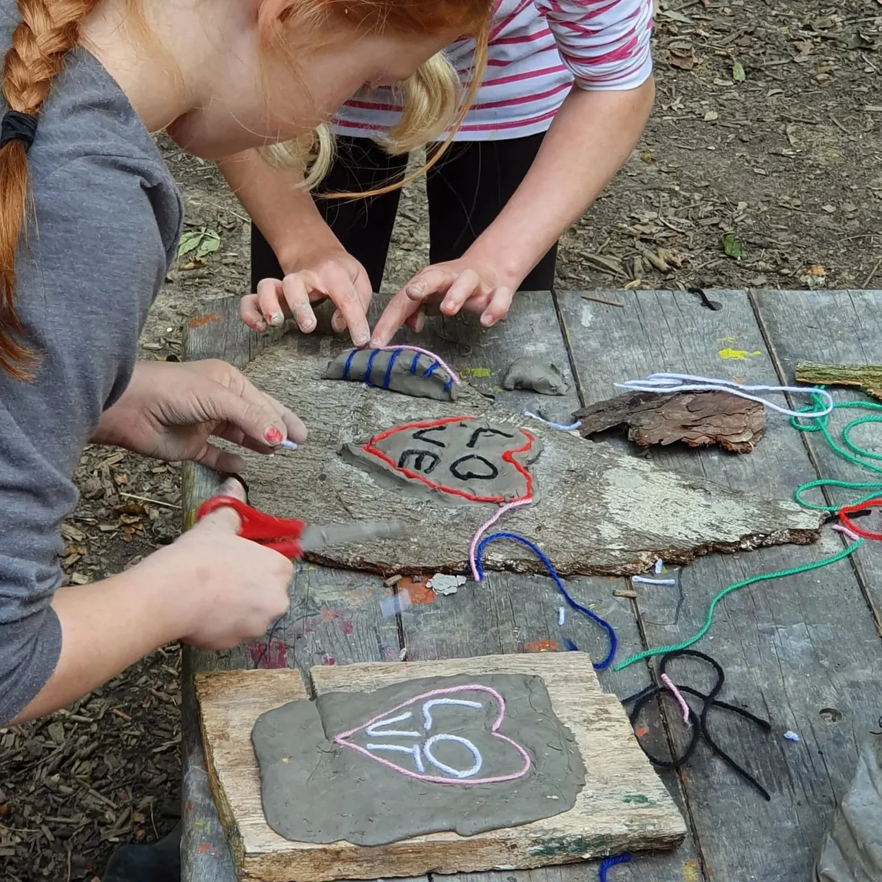 Two children creating nature art on wooden boards, using coloured wires and chalk to draw animal shapes outdoors.