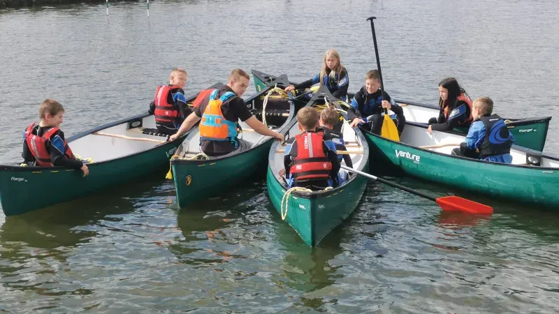 Children in life jackets gather in green canoes on water during a group paddling activity.