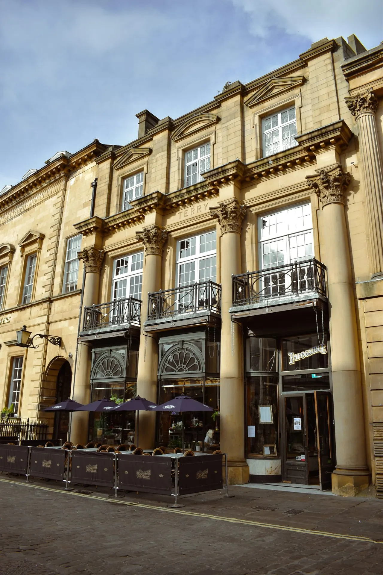 Historic stone building with classical columns and large windows, featuring outdoor seating with purple umbrellas beneath.