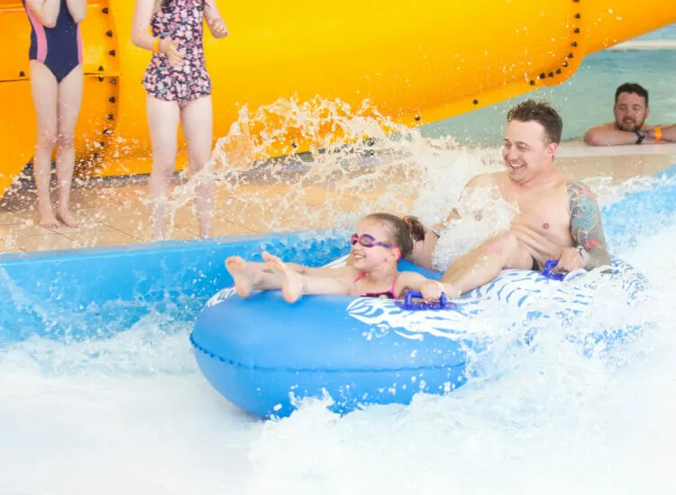 Children and adults enjoying a wave pool ride on inflatable floats at an indoor water park.