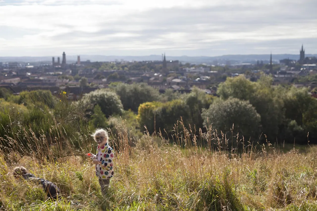 A small child in a spotted top stands in grassland and reeds overlooking a city skyline with church spires and distant hills.