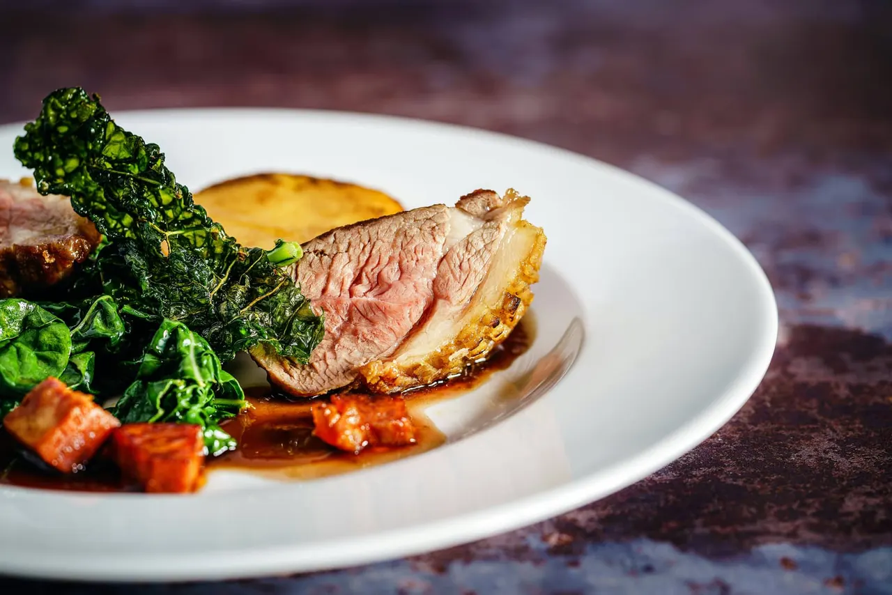 Sliced rare beef with crispy edges, served with roasted vegetables, sautéed greens and sauce on a white plate.