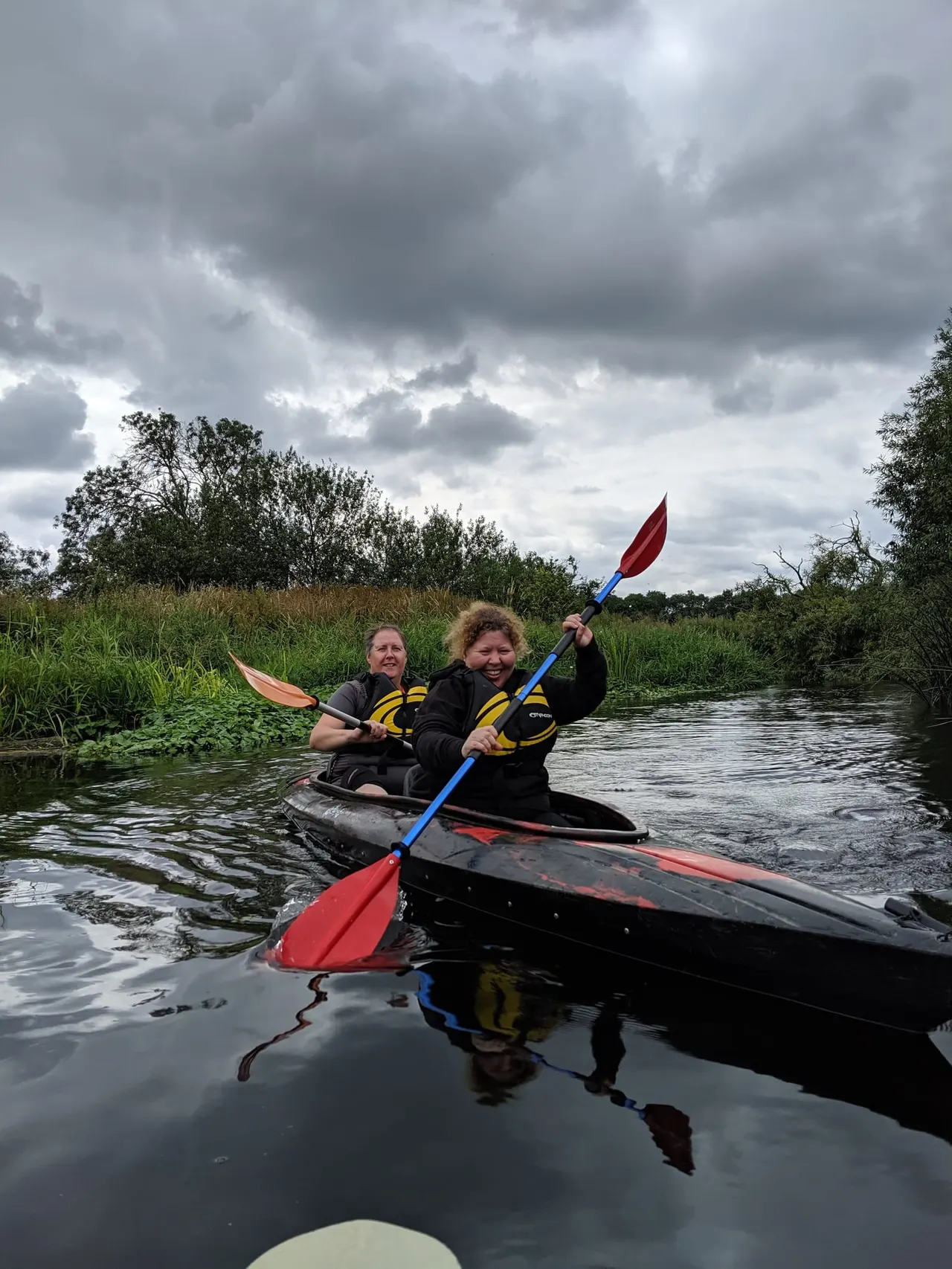 Two people kayaking on a river lined with reeds and trees under a cloudy sky.