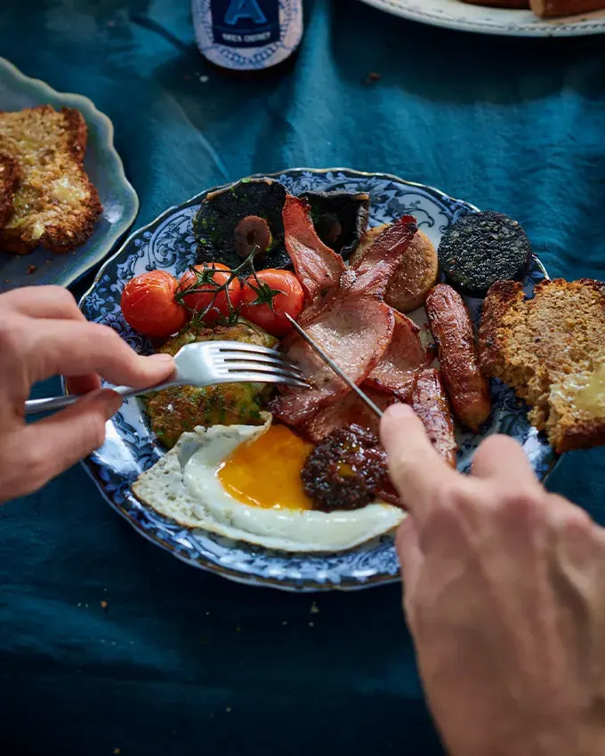 Person eating a full Irish breakfast with bacon, sausage, fried egg, tomatoes and toast on a decorative plate.