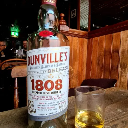 A large bottle of Dunville's 1808 blended Irish whiskey displayed on a wooden bar counter with a glass of whiskey beside it.
