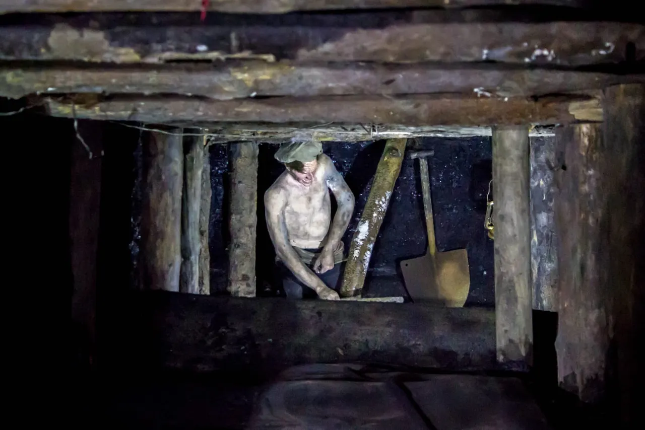 Man crouching in a cramped underground mine tunnel with wooden supports and mining tools.