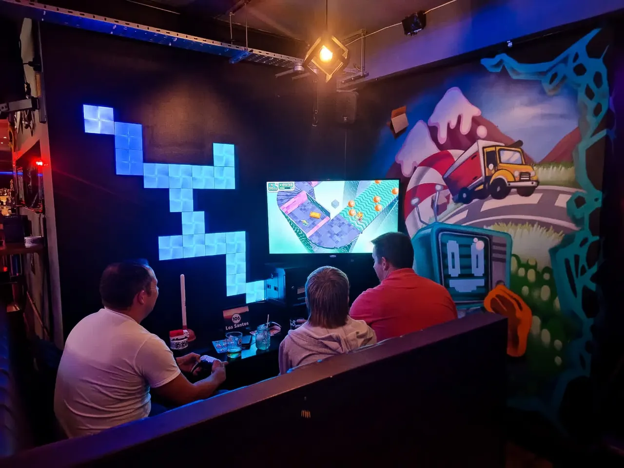 Three people playing retro arcade games at a neon-lit gaming bar with colourful screen displays and themed wall art.