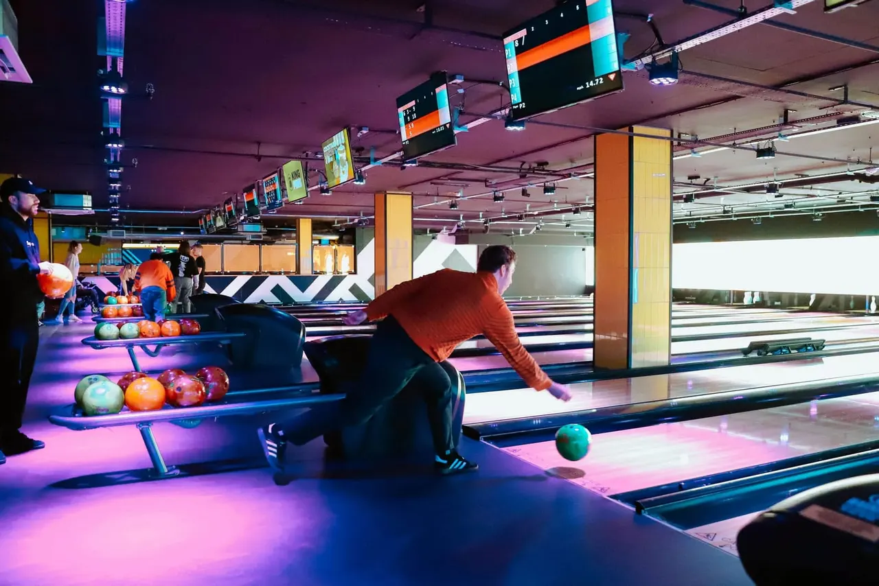 Man in red shirt bowling on neon-lit lane with coloured balls and other bowlers visible in modern bowling alley.