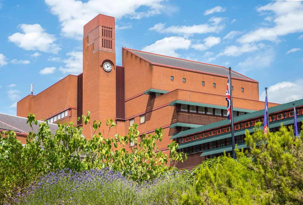 Modern red-brick building with clock tower and green balconies, surrounded by lavender and shrubs under blue sky.