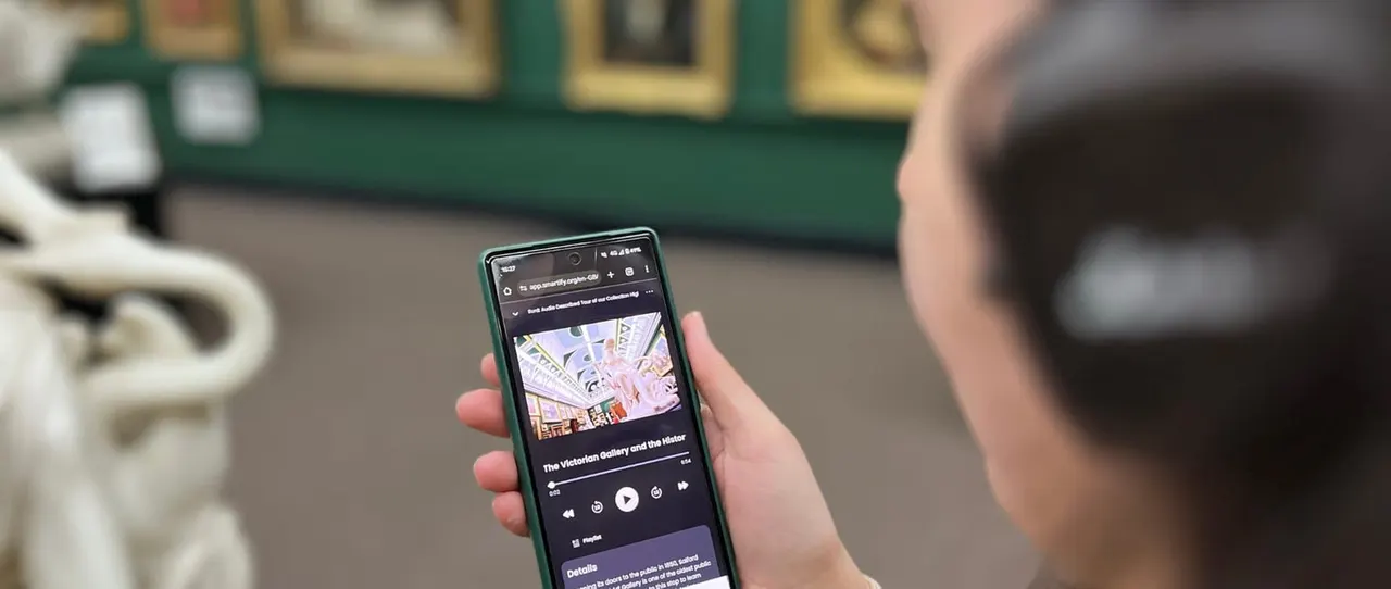 Person holding a smartphone displaying an audio guide for The Victorian Gallery and the Historic, with a museum building visible blurred in the background.