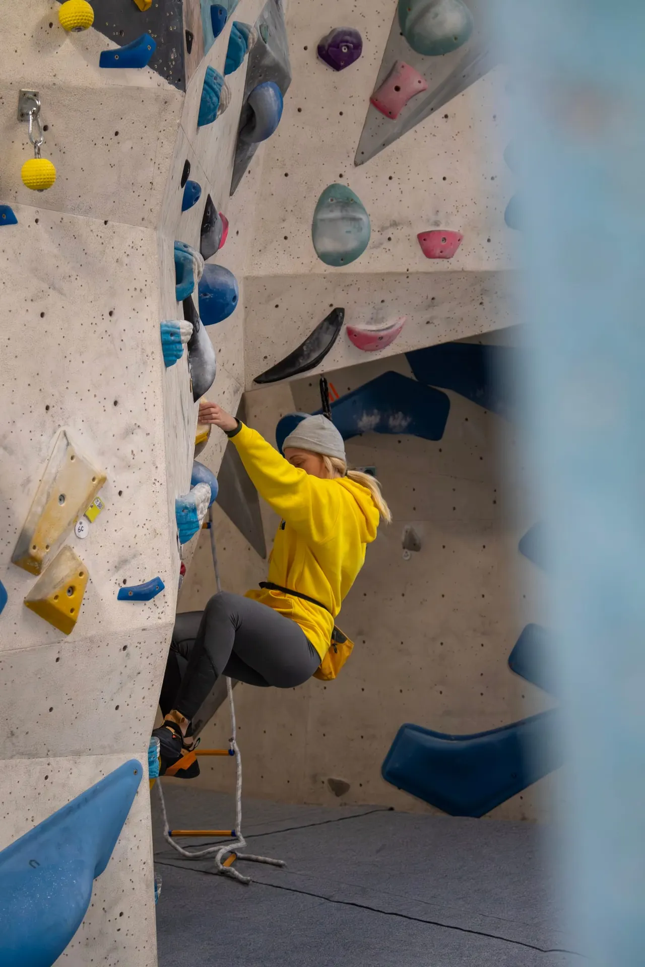 Person in yellow jacket climbing an indoor rock wall with coloured handholds and protective rope.
