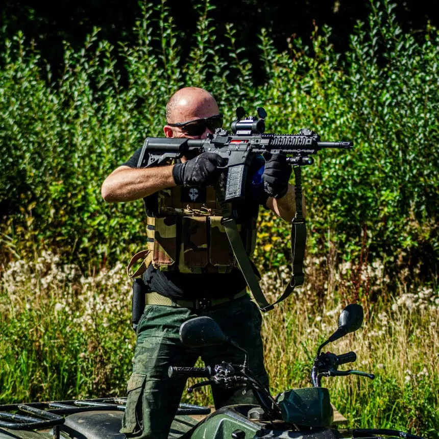 Man kneeling on ground aiming airsoft rifle at outdoor range with vegetation backdrop.