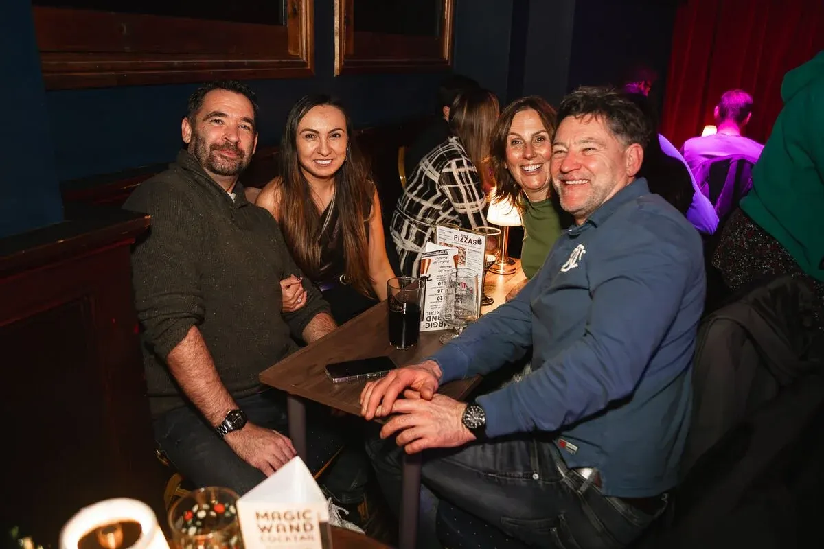 Five people smiling at a table with drinks in an indoor venue with coloured lighting.