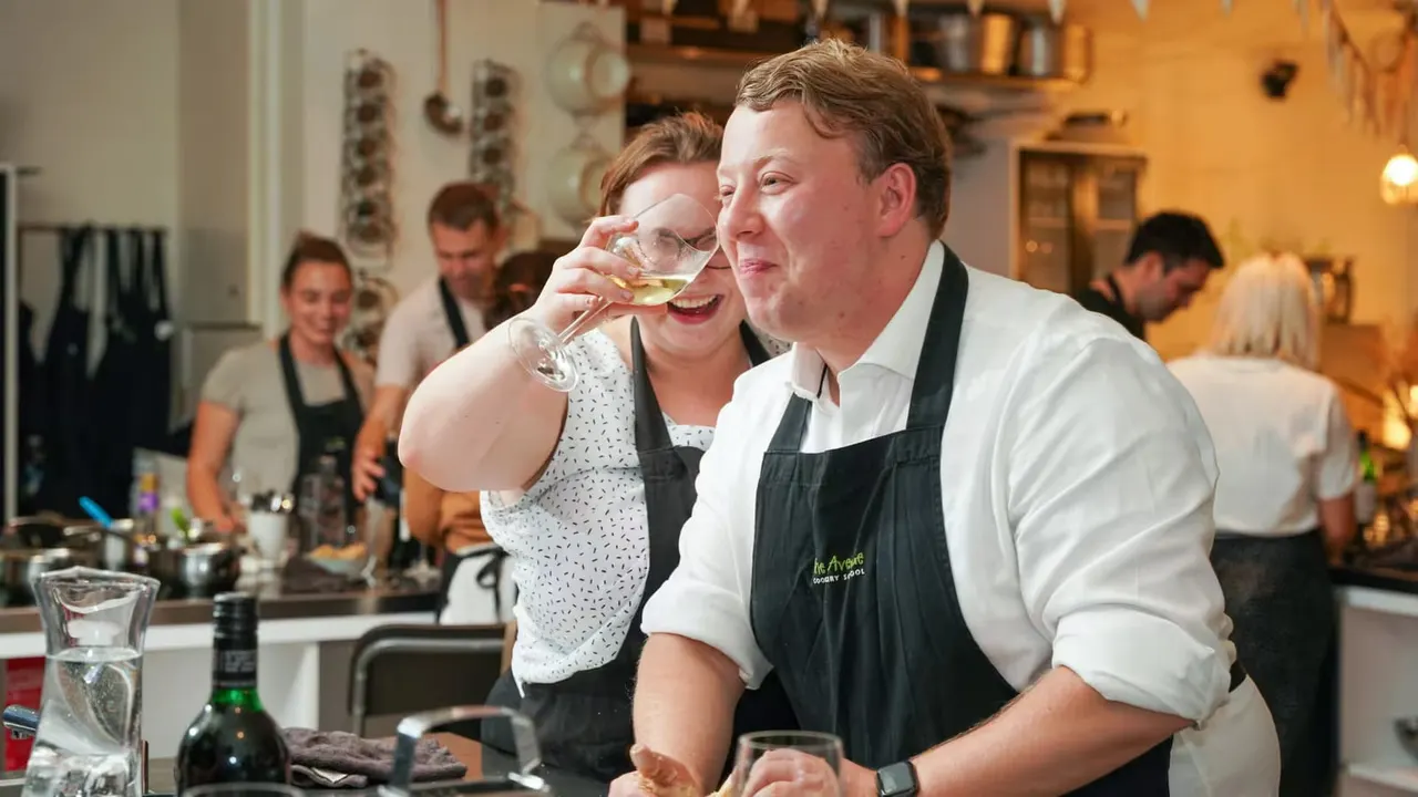 Two staff members in aprons taste wine at a kitchen counter during a cookery class, with other participants visible in the background.