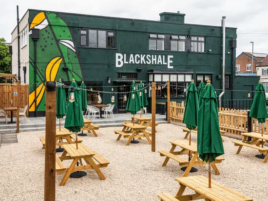 Outdoor seating area with wooden picnic tables under green umbrellas in front of a green-painted building.