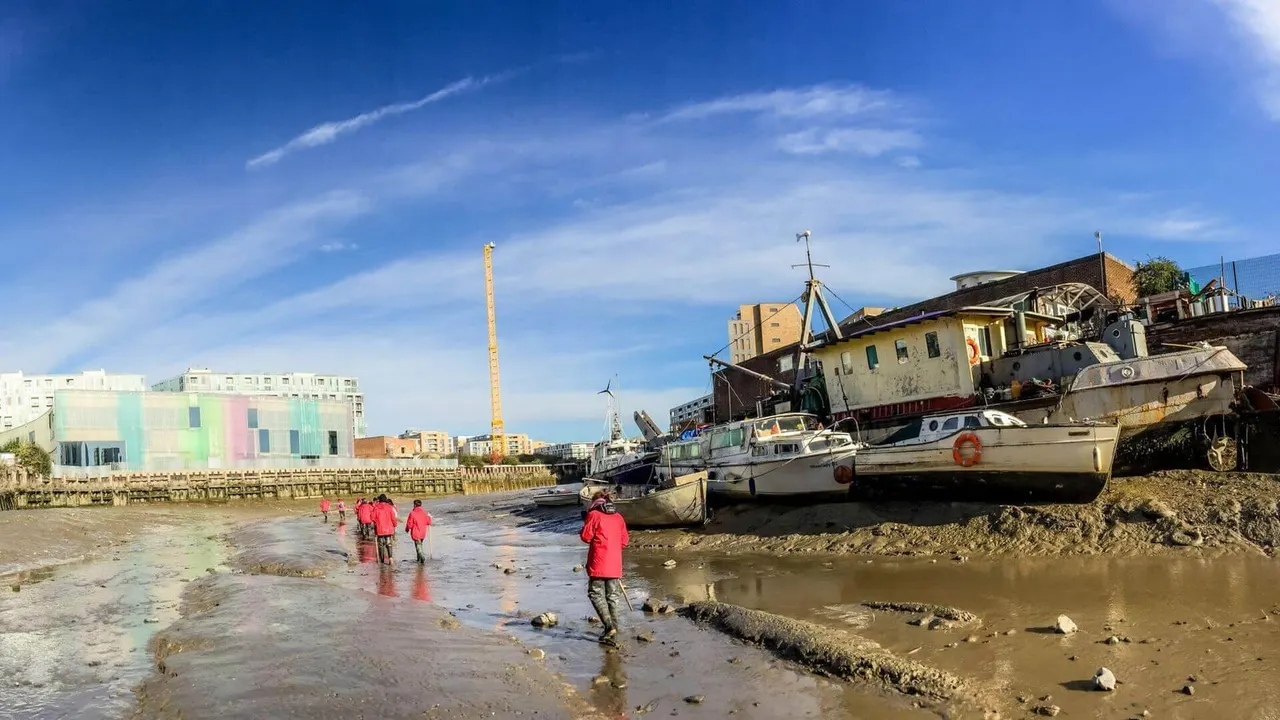 People in red jackets wading in muddy tidal waters amongst moored boats and historic buildings along a riverbank.