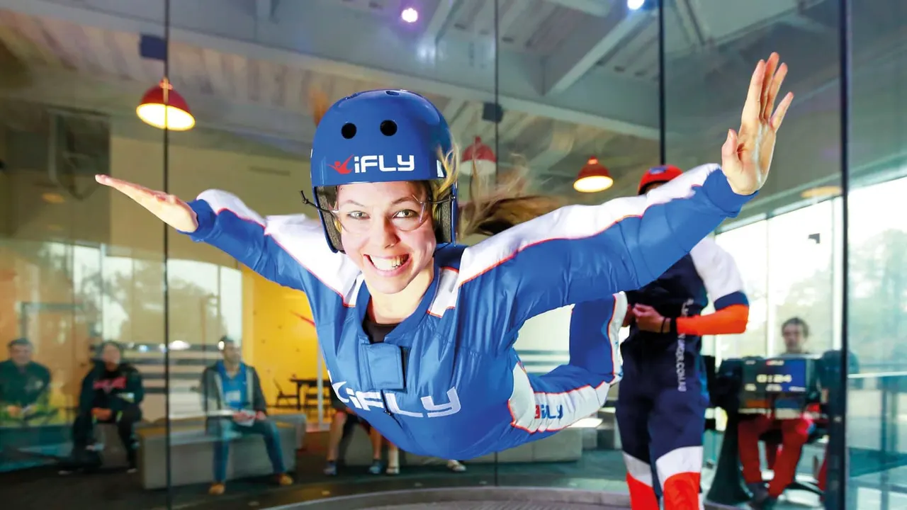 Young person in blue iFLY suit and helmet performing indoor skydiving with arms outstretched, watched by spectators behind glass.