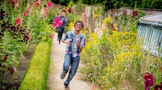 A young boy runs along a garden path lined with colourful flowering plants, with other visitors walking behind him.