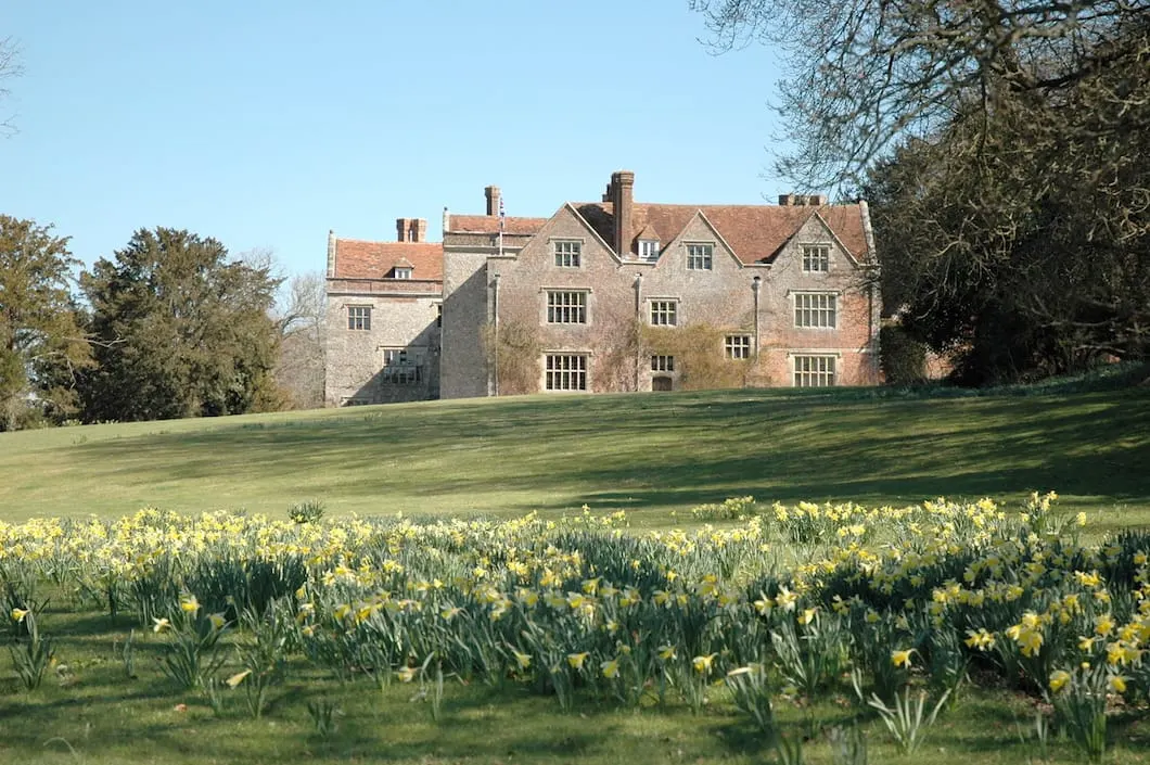 Historic brick manor house set behind a sloping lawn dotted with yellow daffodils in spring.