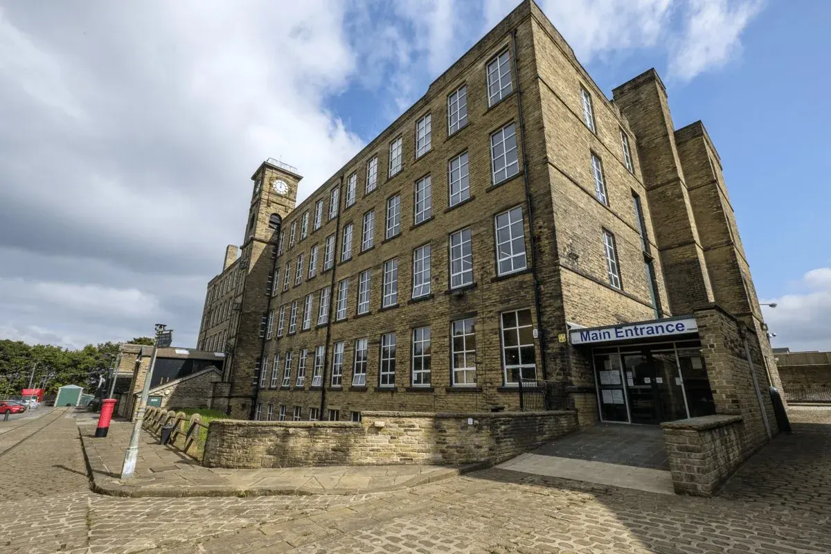Large stone mill building with clock tower and "Main Entrance" sign, set in paved courtyard under partly cloudy sky.
