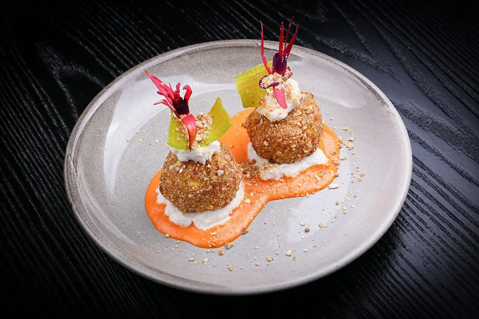 Two golden croquettes with white cream, green fruit, and red flower garnish on orange sauce on a grey plate.